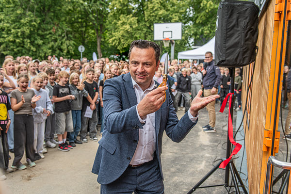 Borgmesteren klipper den røde snor ved indvielsen af den nye skolebygning, mens en stor gruppe børn og voksne ser til i baggrunden.